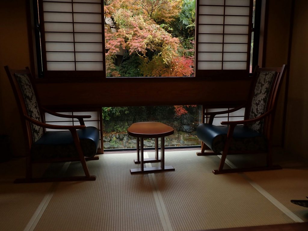 Interior de ryokan tradicional en Japón con tatami, sillas bajas y ventana con jardín otoñal.